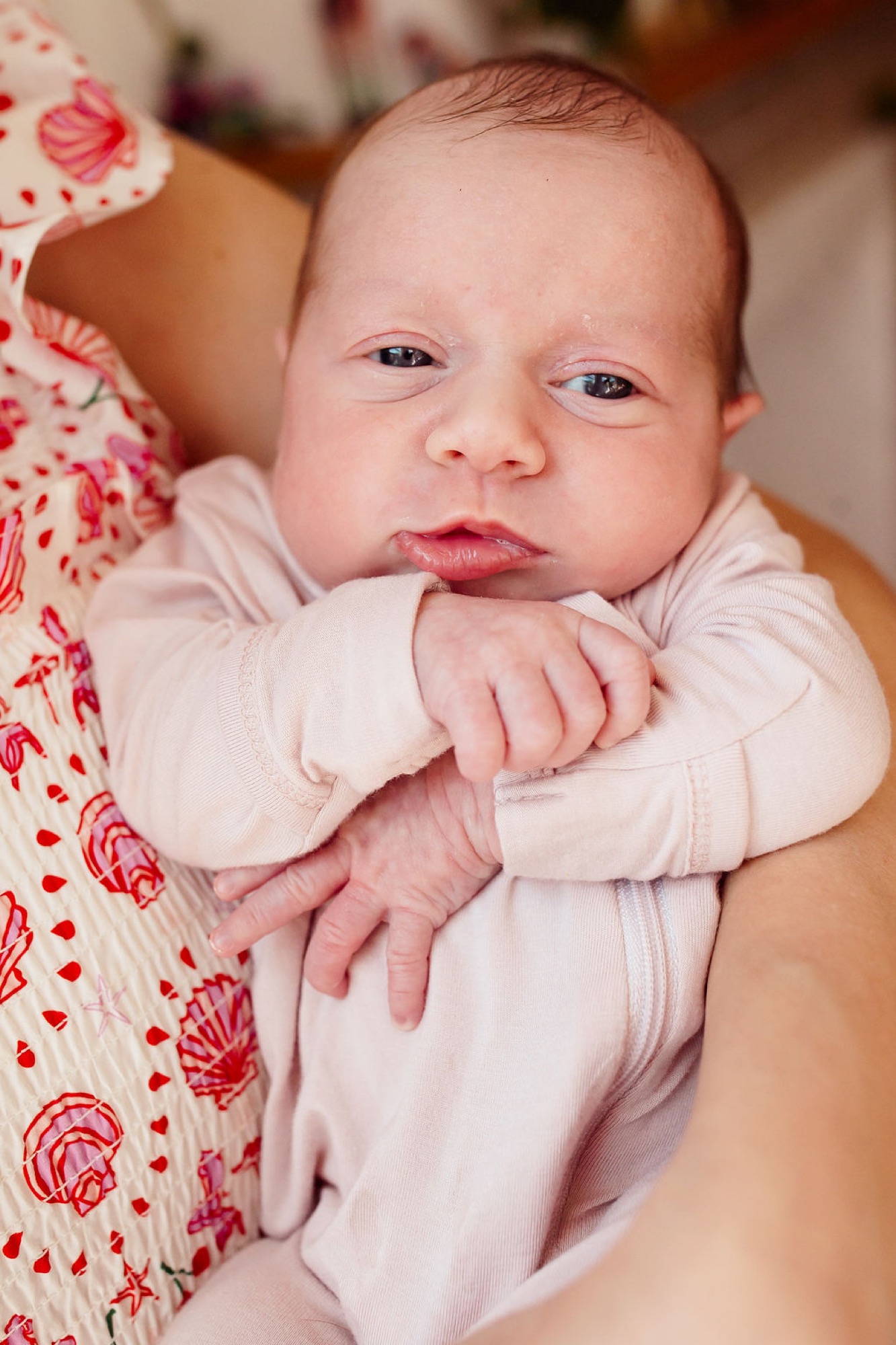 Newborn in pale pink outfit rests against parent's red floral clothing, gazing directly at camera with alert eyes