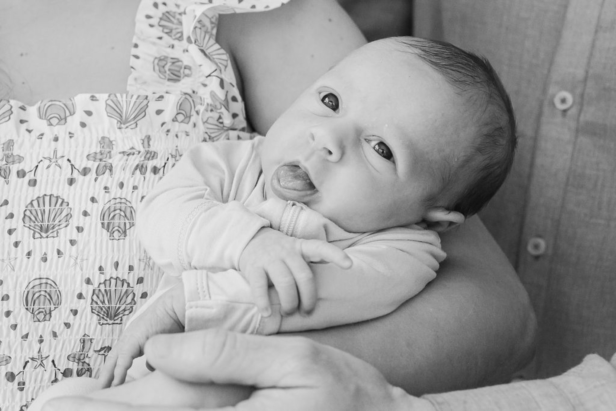 Newborn gazes upward while cradled against a parent's bare chest in soft natural light, tiny fist grasping fabric