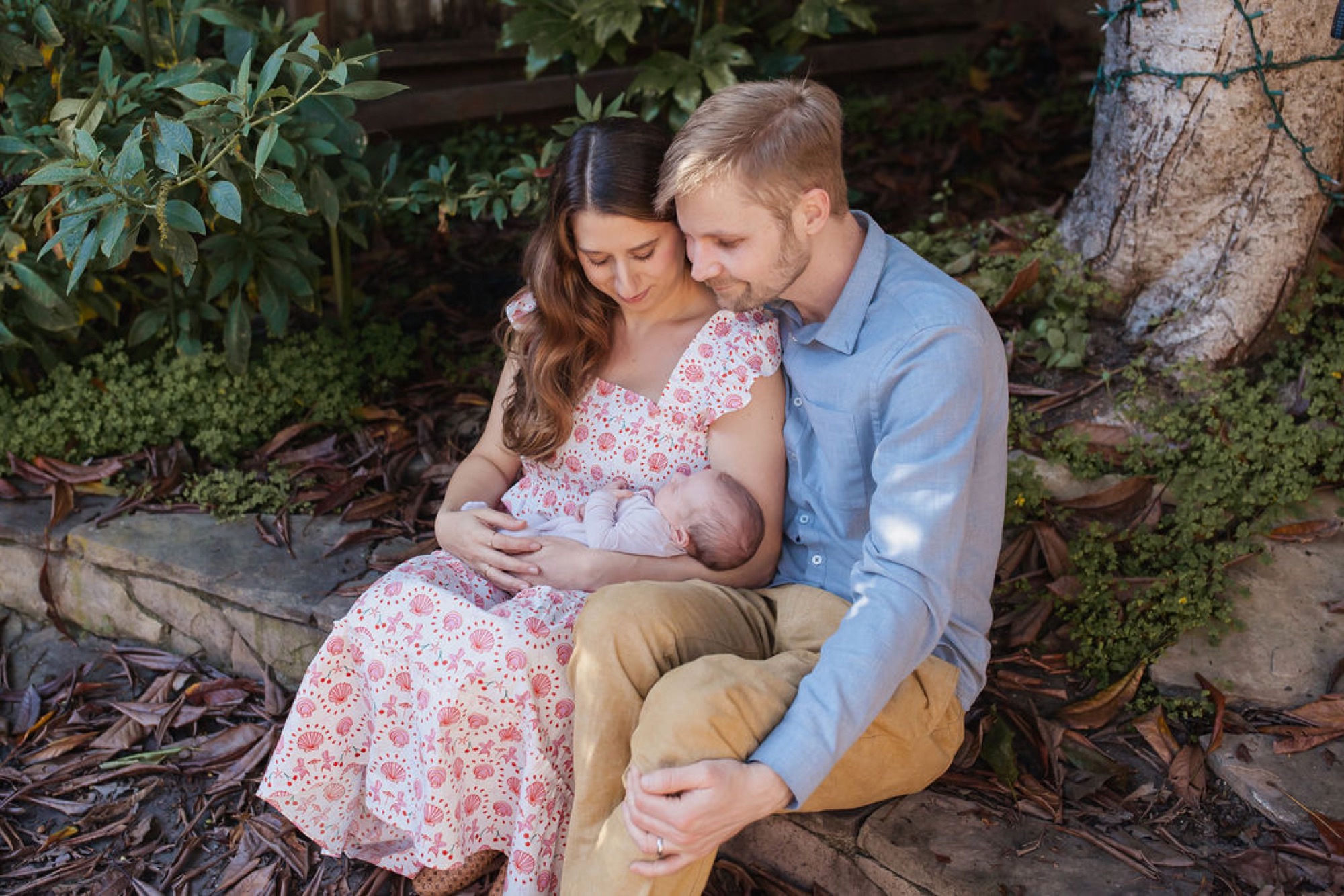 Parents sit close on a garden log, tenderly cradling their newborn while surrounded by lush green foliage and dappled natural light