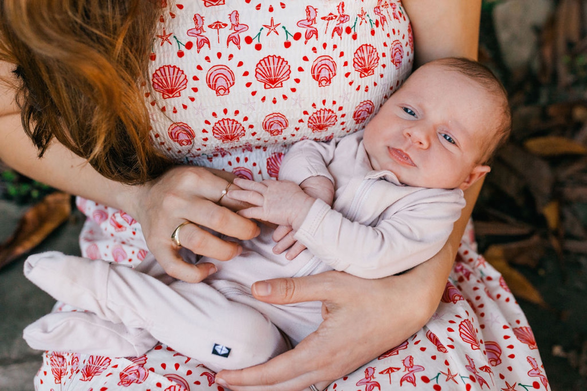 Mother in red shell-patterned dress cradles newborn in white, their hands gently touching as soft indoor light illuminates the tender moment