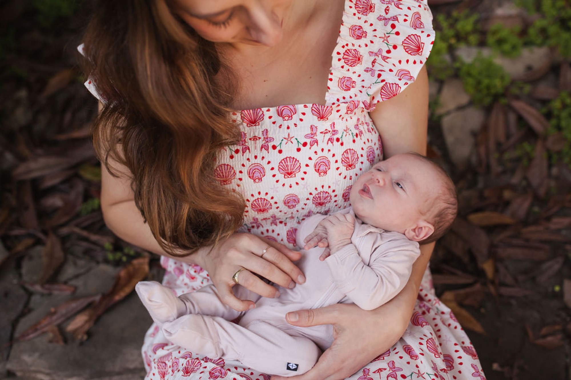 Mother in pink floral dress cradles sleeping newborn against her chest in dappled woodland light