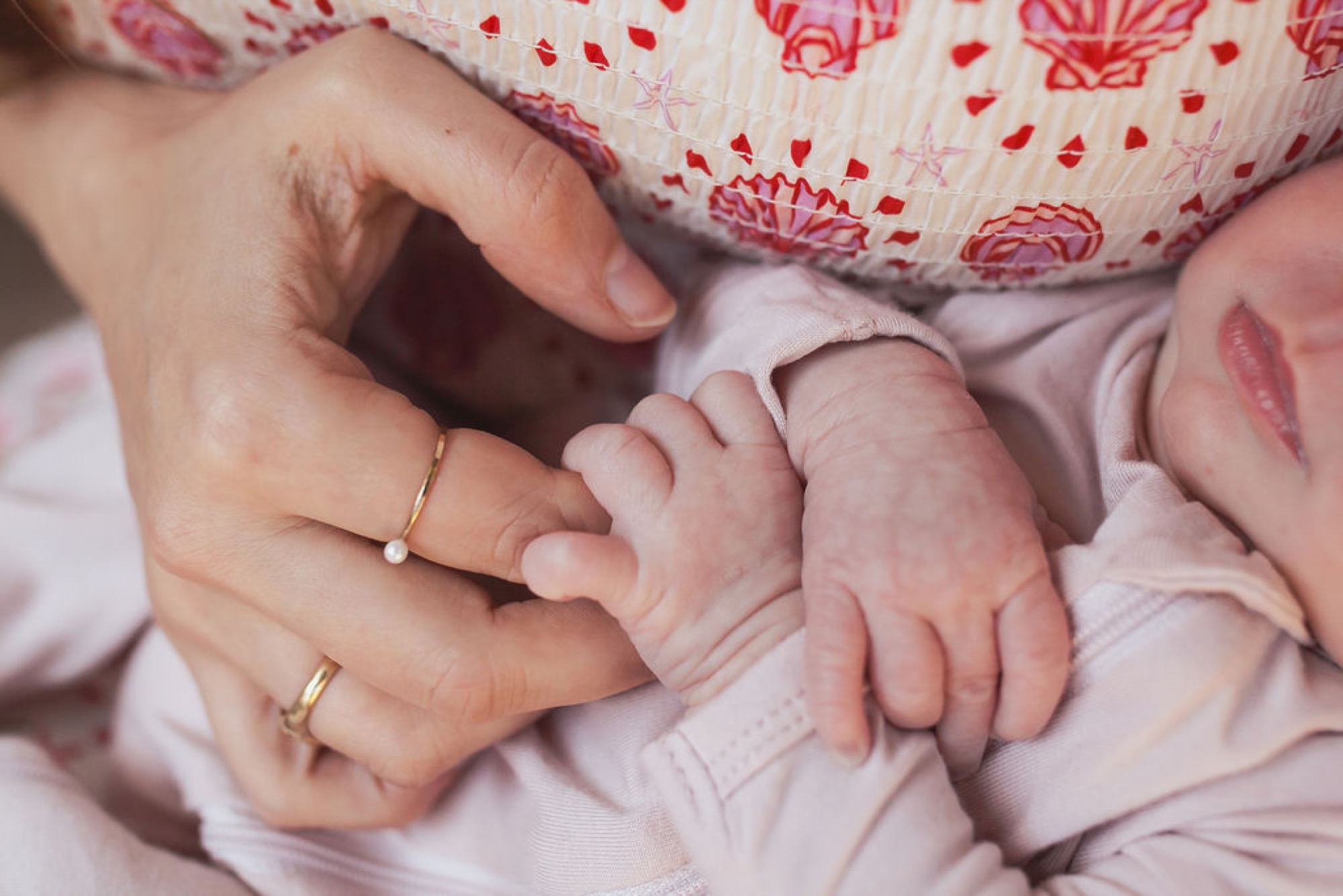 A mother's hands gently cradle her newborn's tiny fingers and palms in soft natural light during an intimate indoor portrait session