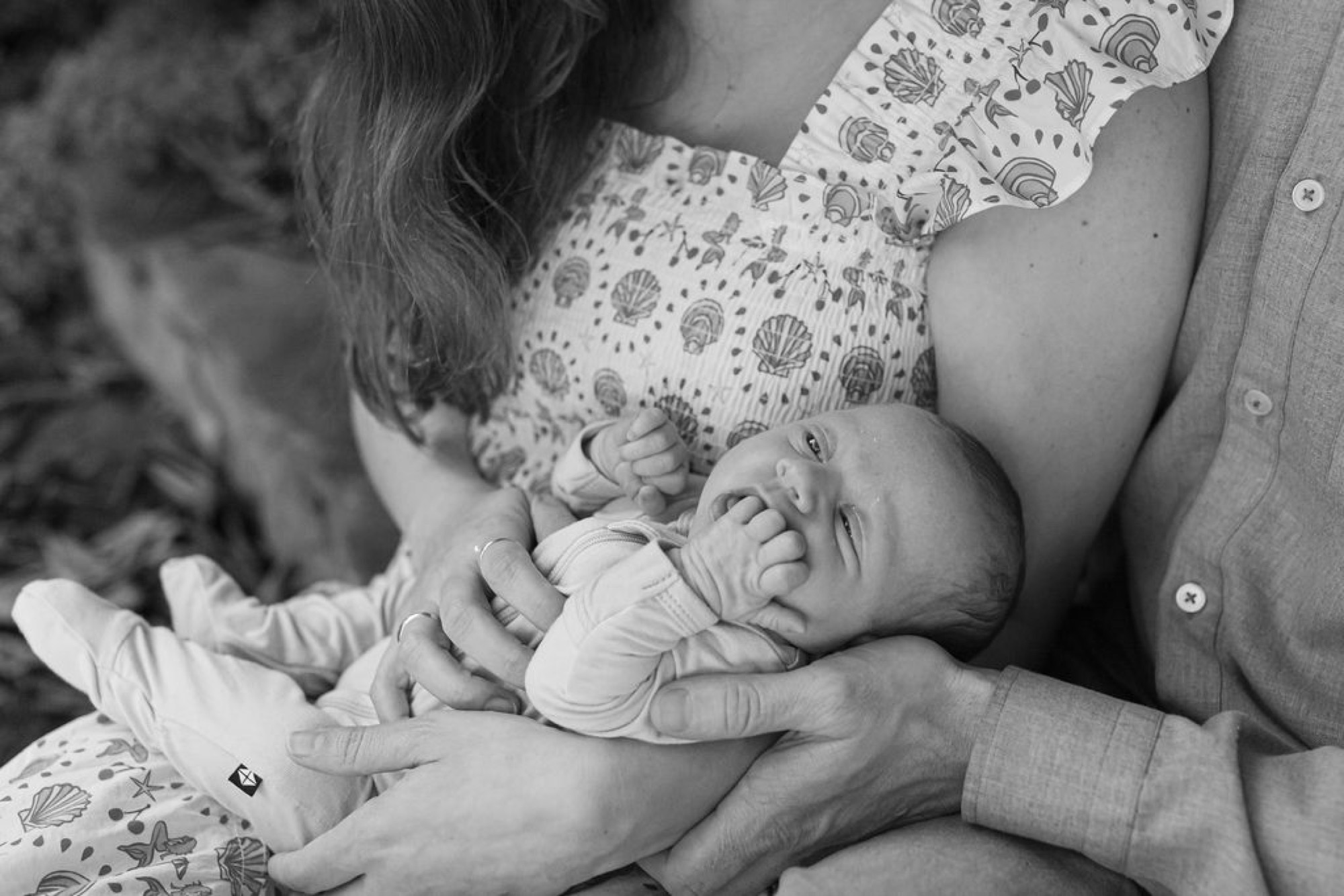 A newborn sleeps peacefully in a parent's arms while an older sibling leans in close, rendered in soft black and white