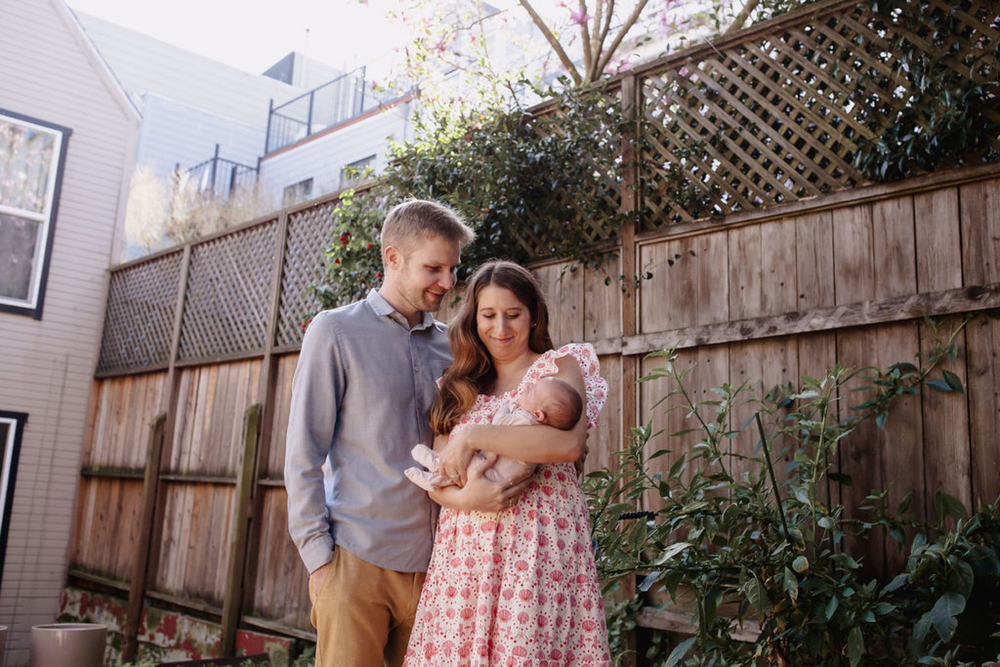 Parents gaze down at their newborn in a sunny backyard, wooden fences and climbing vines framing the intimate moment