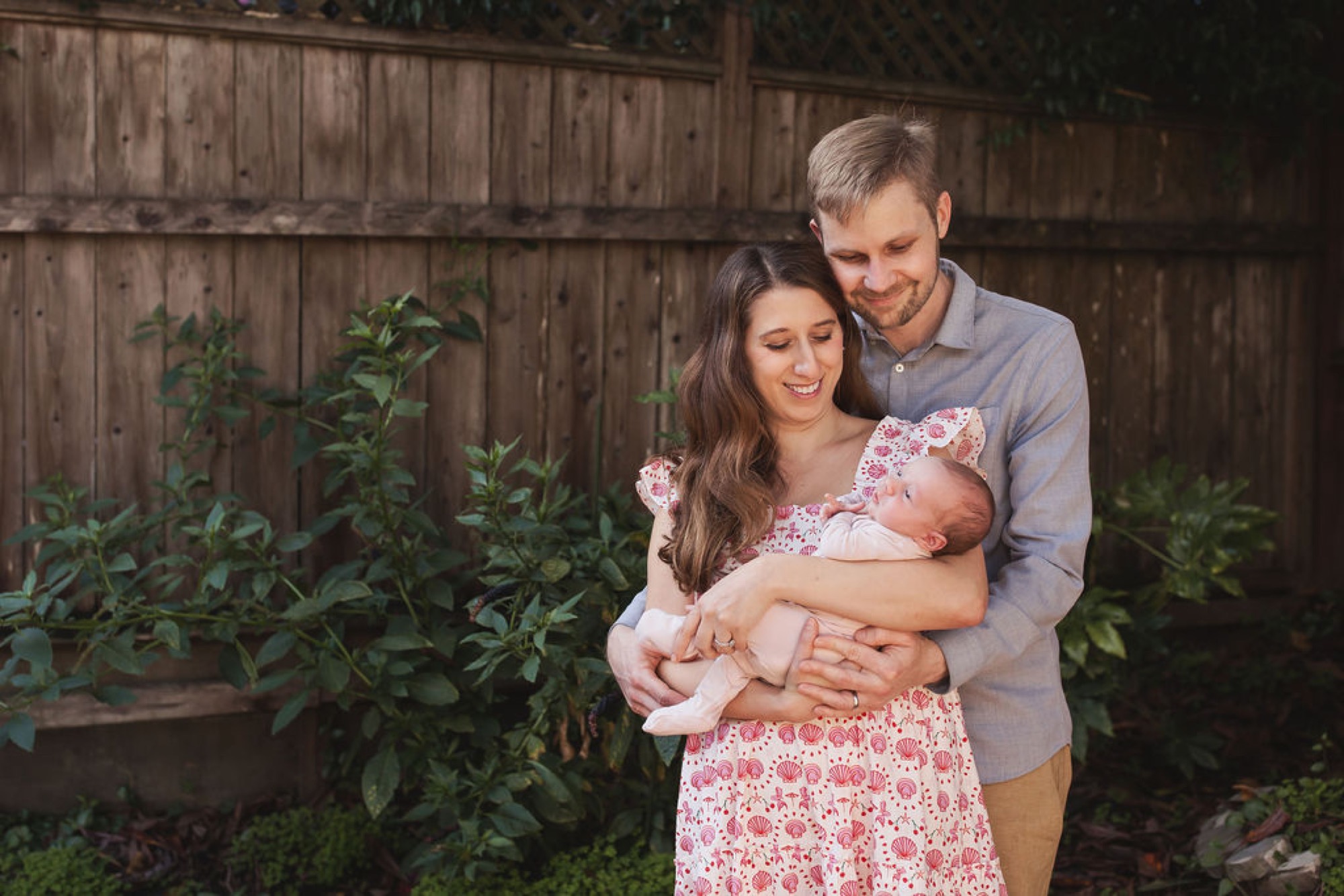 Parents beam down at their newborn in a wooden-fenced garden, soft afternoon light catching the mother's floral dress and baby's delicate features