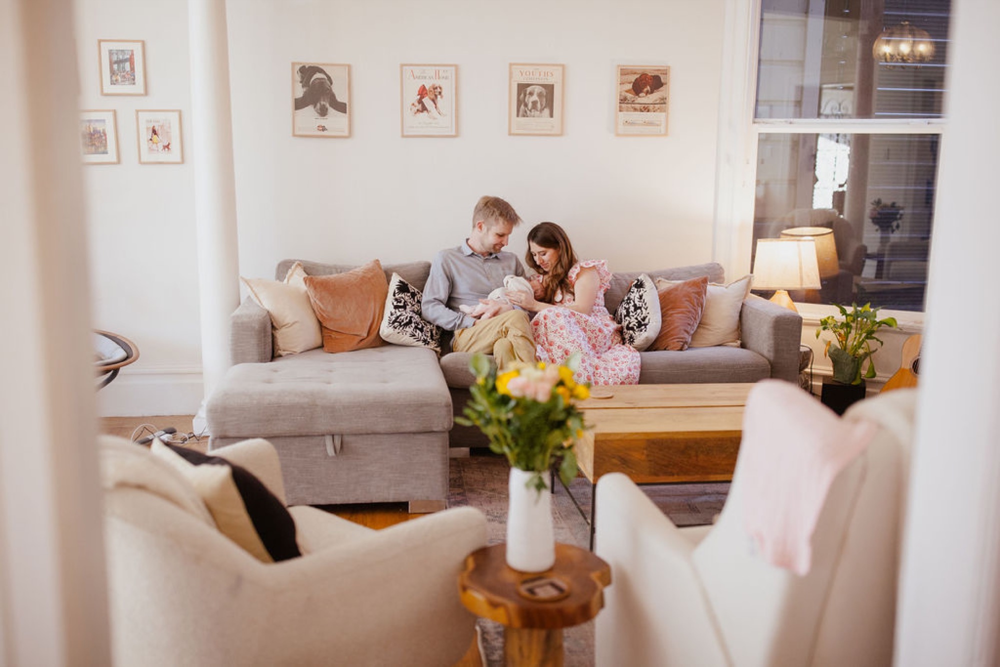 Parents sit close on a beige sofa, gazing down at their newborn swaddled in pink, while warm afternoon light filters through windows behind them