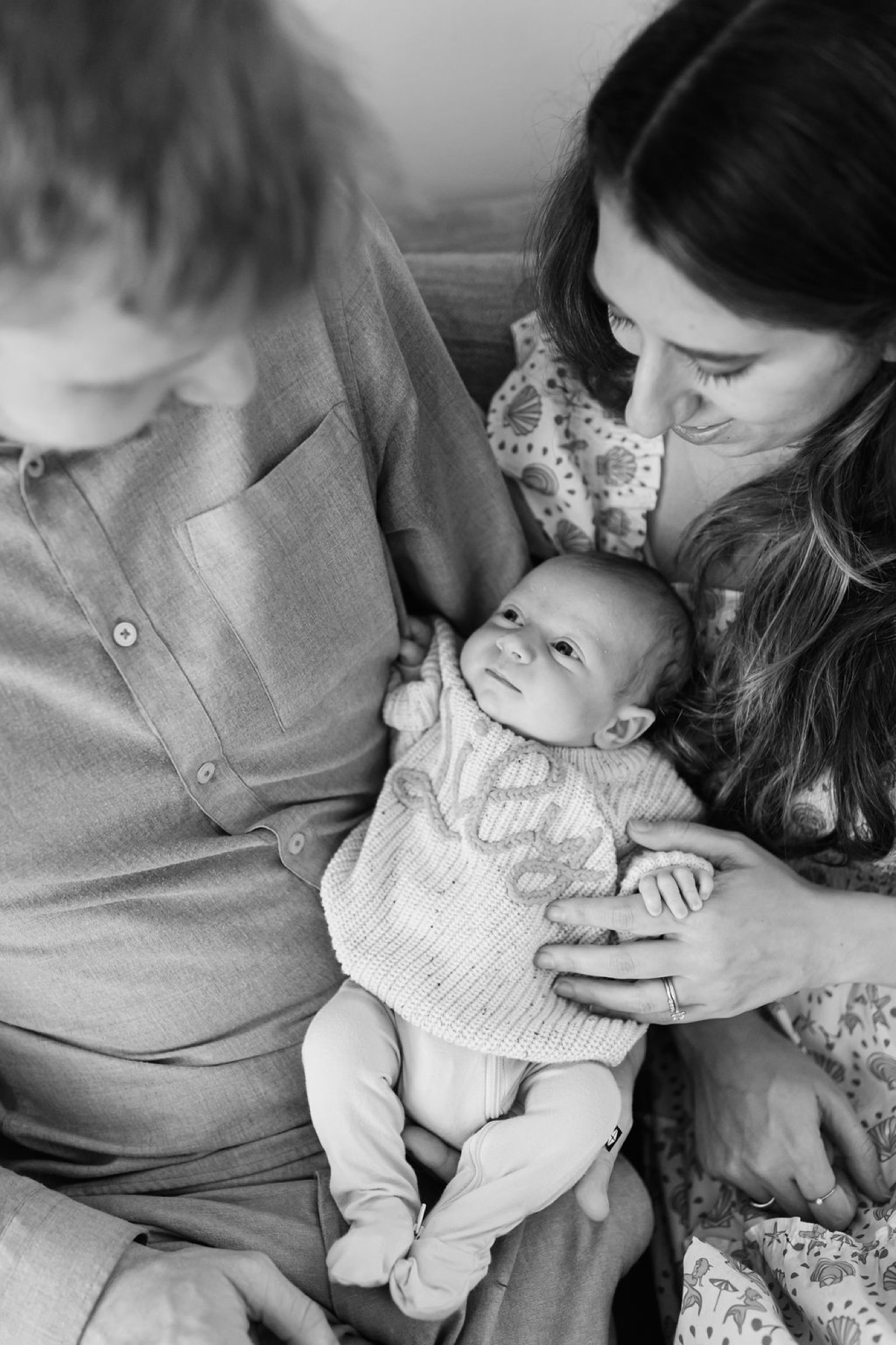 Two parents gaze down at their newborn cradled between them in black and white, tender expressions framing the infant's peaceful face