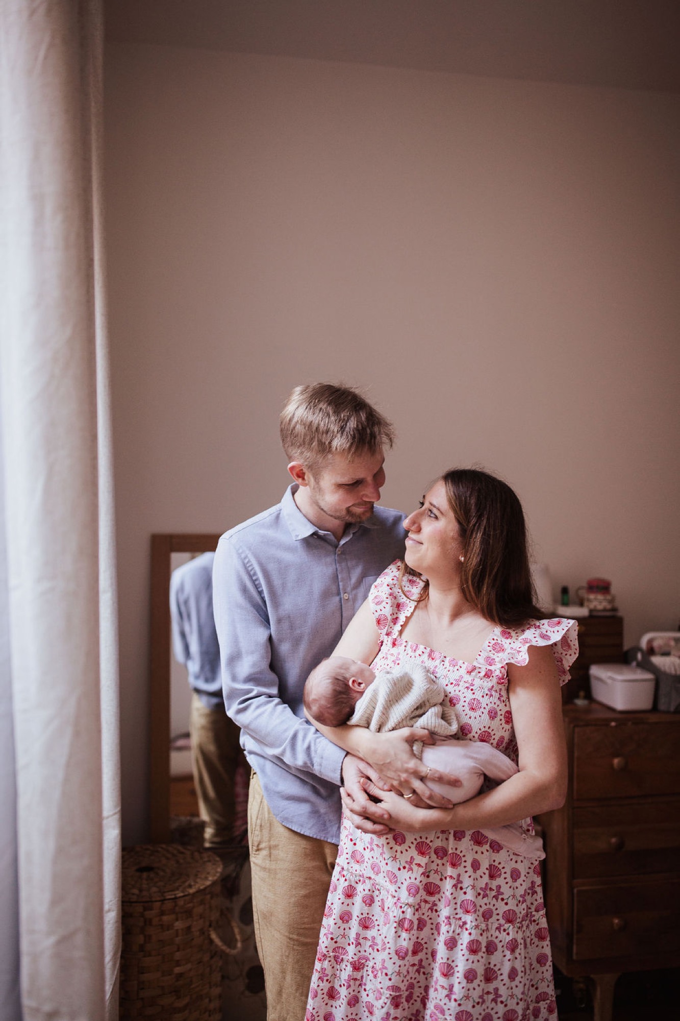 Parents gaze tenderly at their newborn in a soft-lit bedroom, the father in blue shirt and mother in pink floral dress cradling their infant together