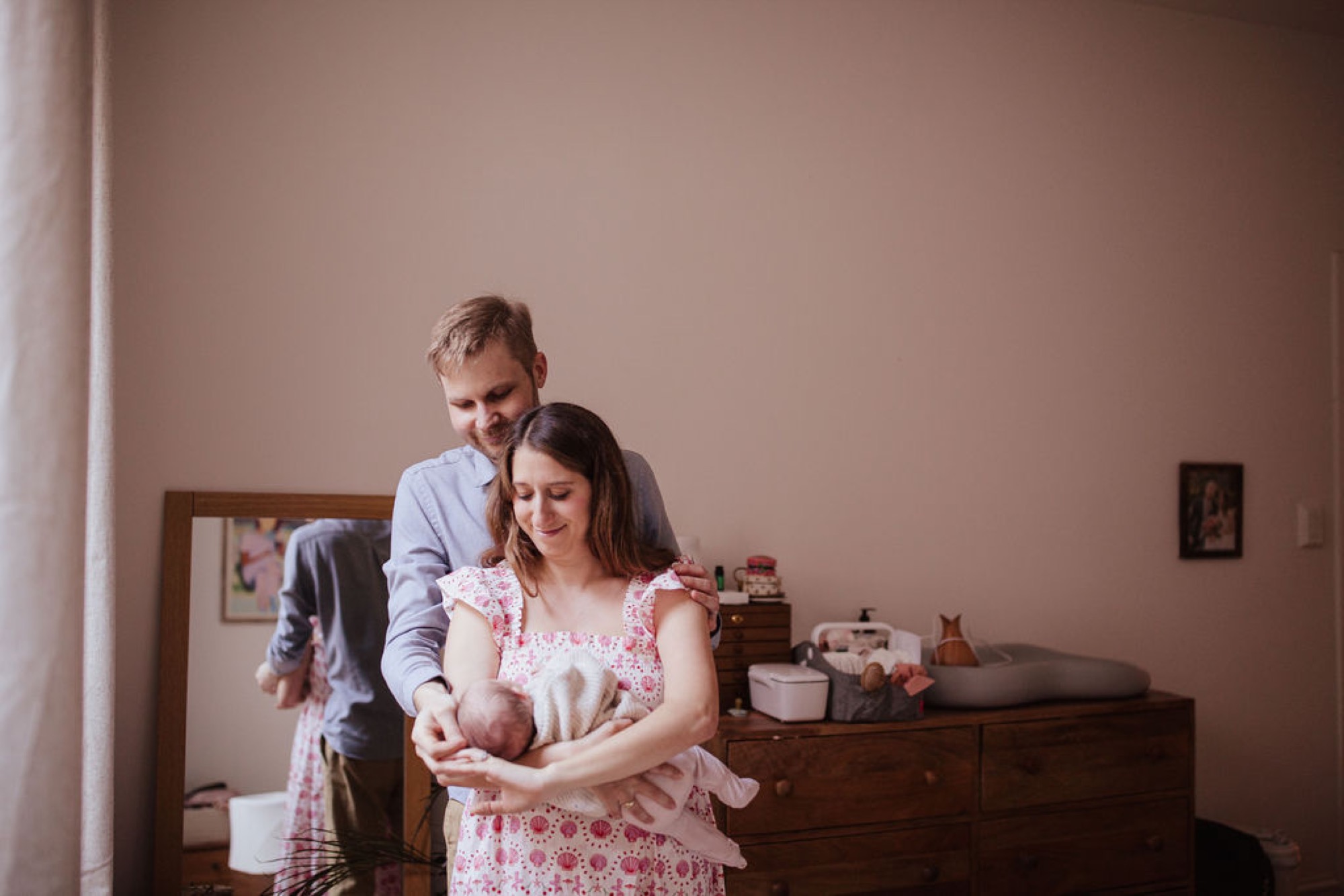 Father stands behind mother cradling their newborn in a soft-lit bedroom, both gazing down with tender expressions at their infant