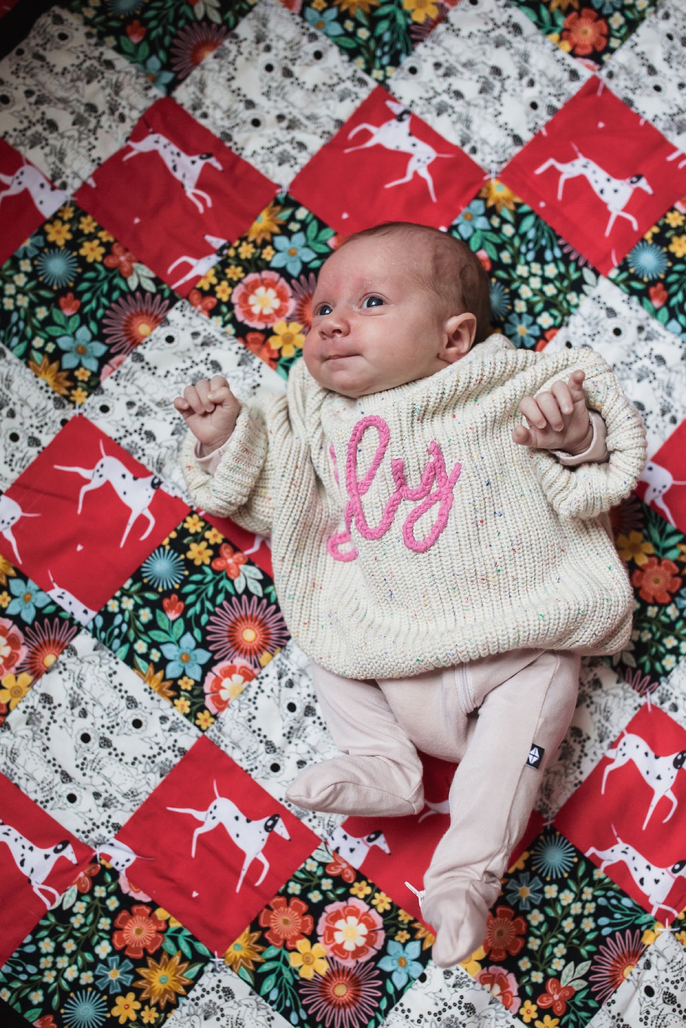 Newborn in cream knit sweater with pink embroidery lies on a patchwork quilt, gazing upward with arms relaxed at sides