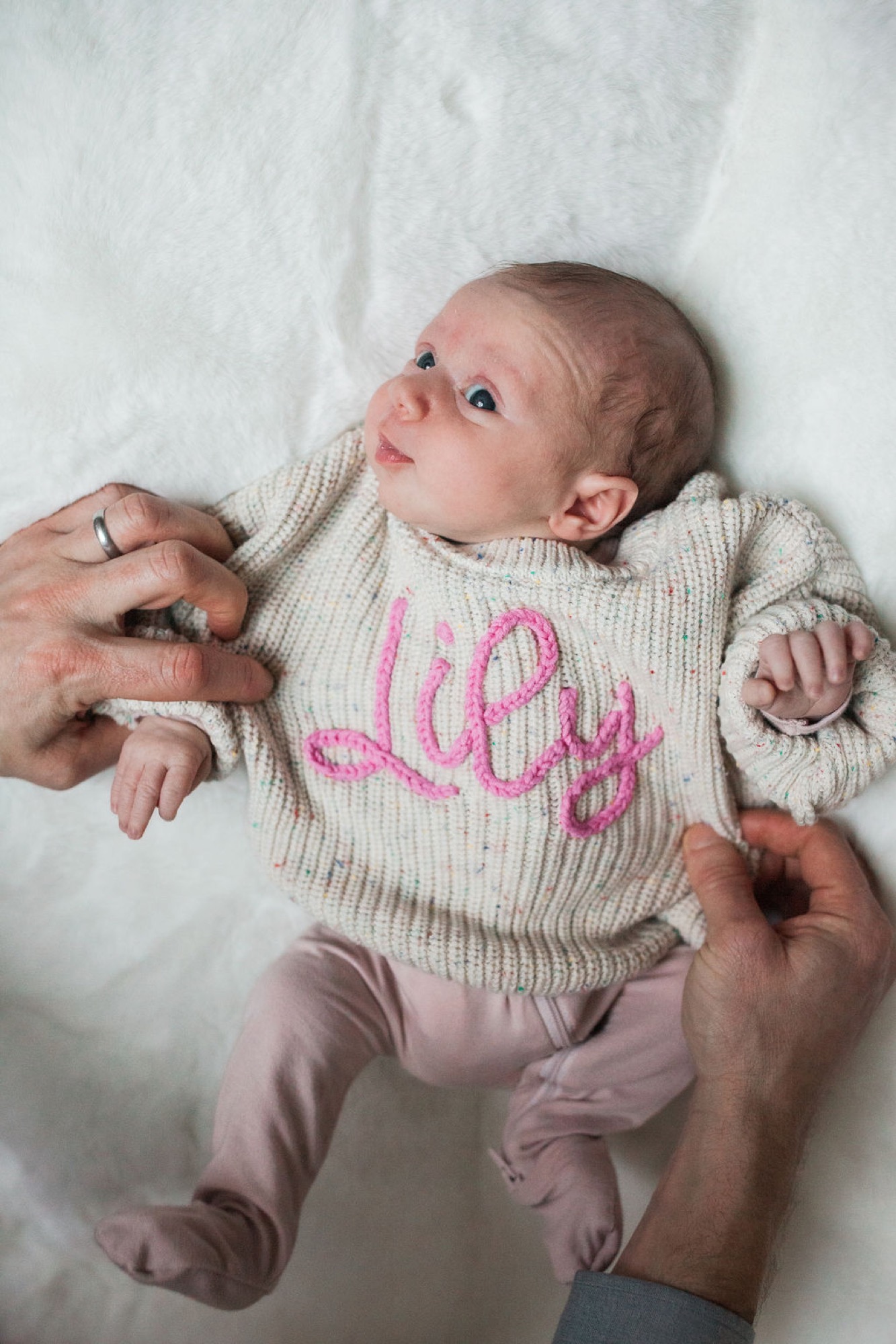 Newborn in cream ribbed sweater with pink embroidered name gazes upward while parents' hands gently support her on white fabric