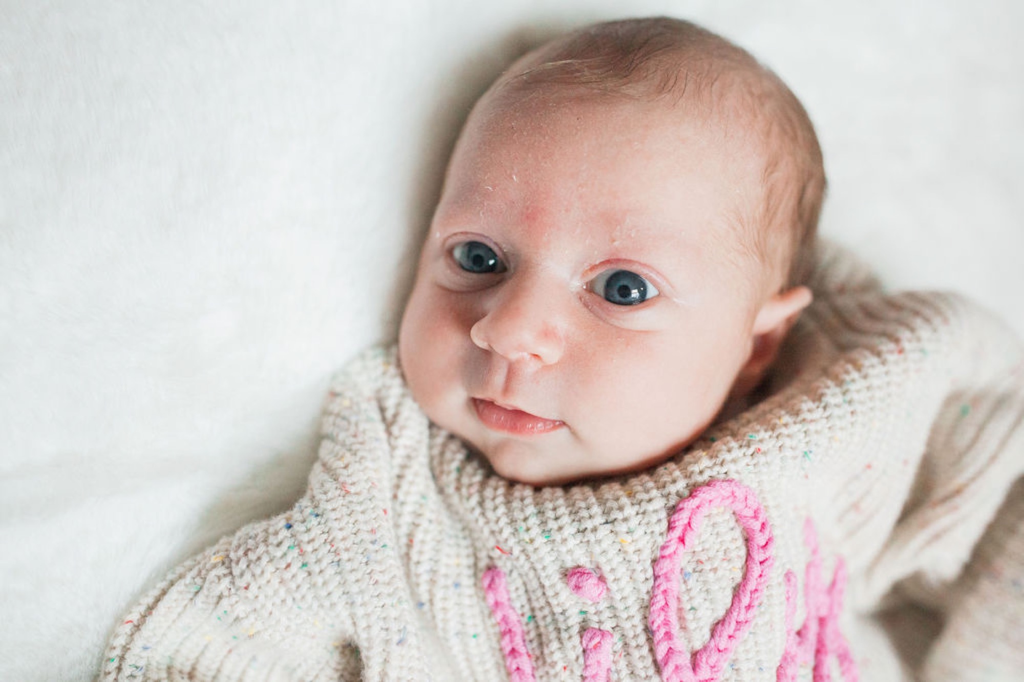 Wide-eyed newborn in cream knit sweater gazes directly at camera with alert expression against white background