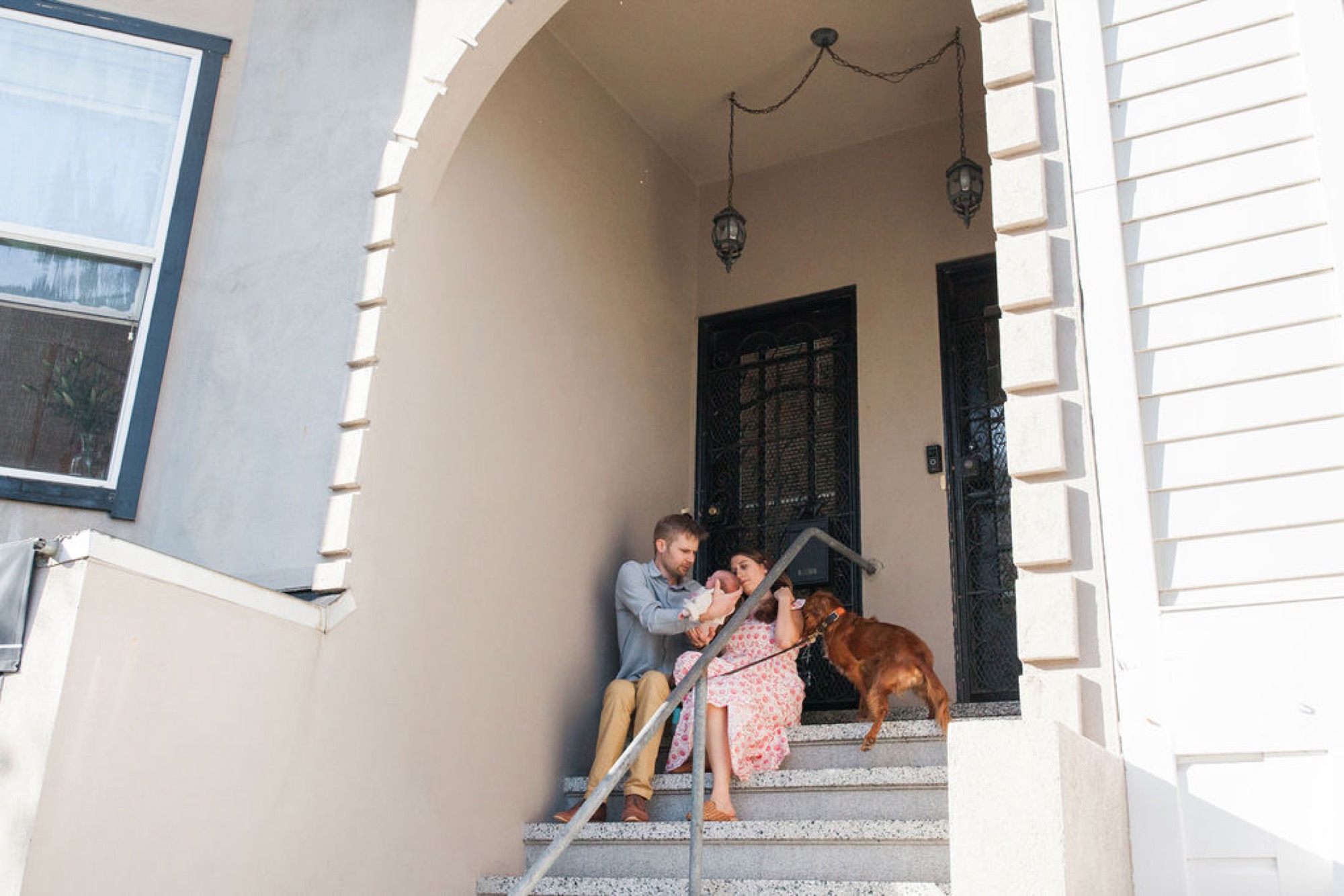 Family sits together on front steps in warm afternoon light, father holding infant while mother and older child lean in close