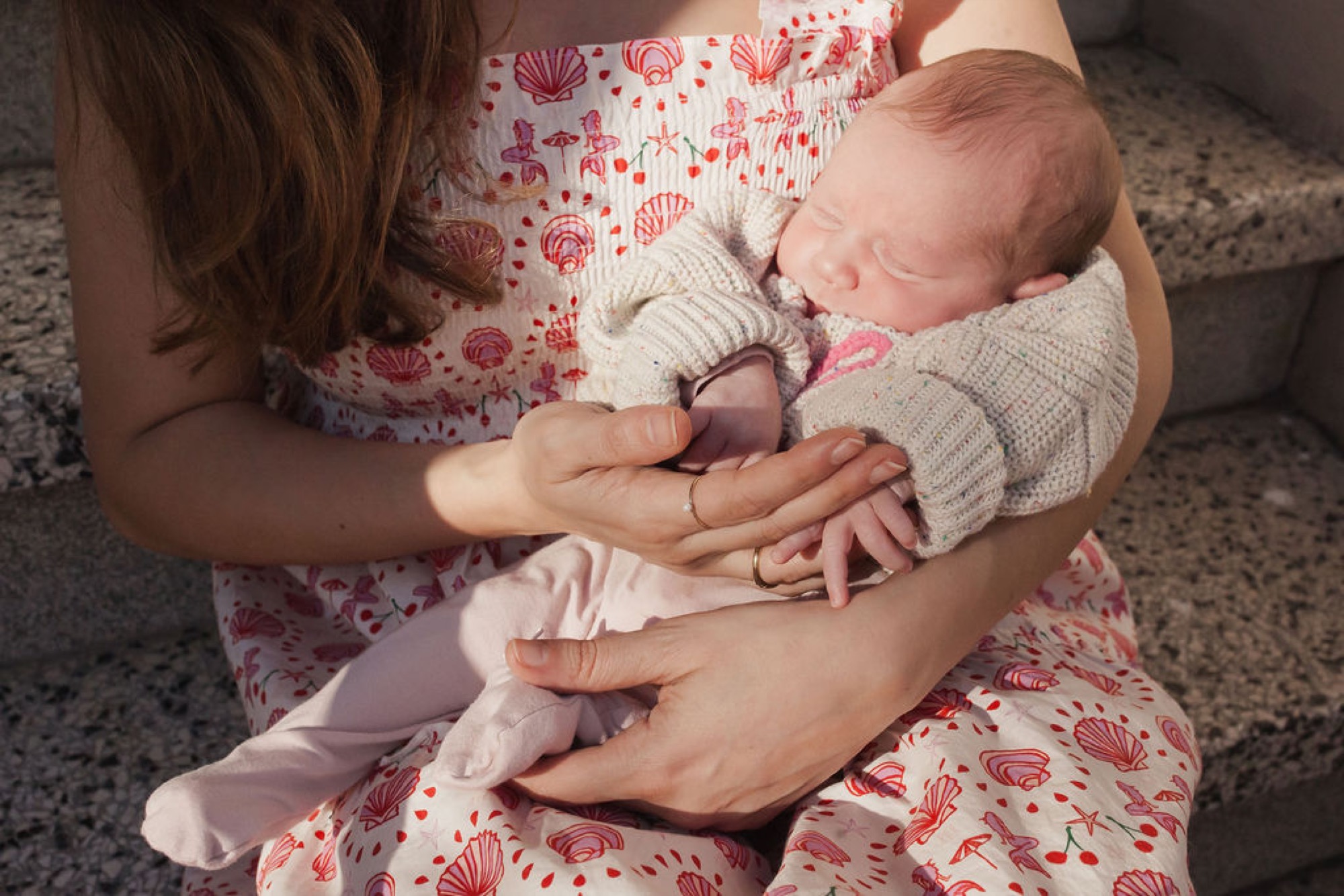 A newborn swaddled in cream knit rests peacefully in a caregiver's hands against a floral-patterned dress on soft carpet