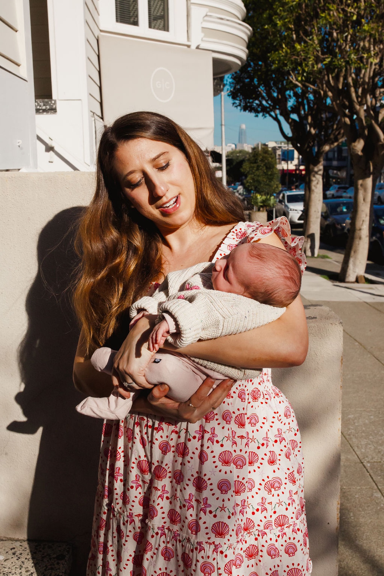 Mother gazes down at her newborn wrapped in white, bathed in warm sunlight on a residential sidewalk with trees behind