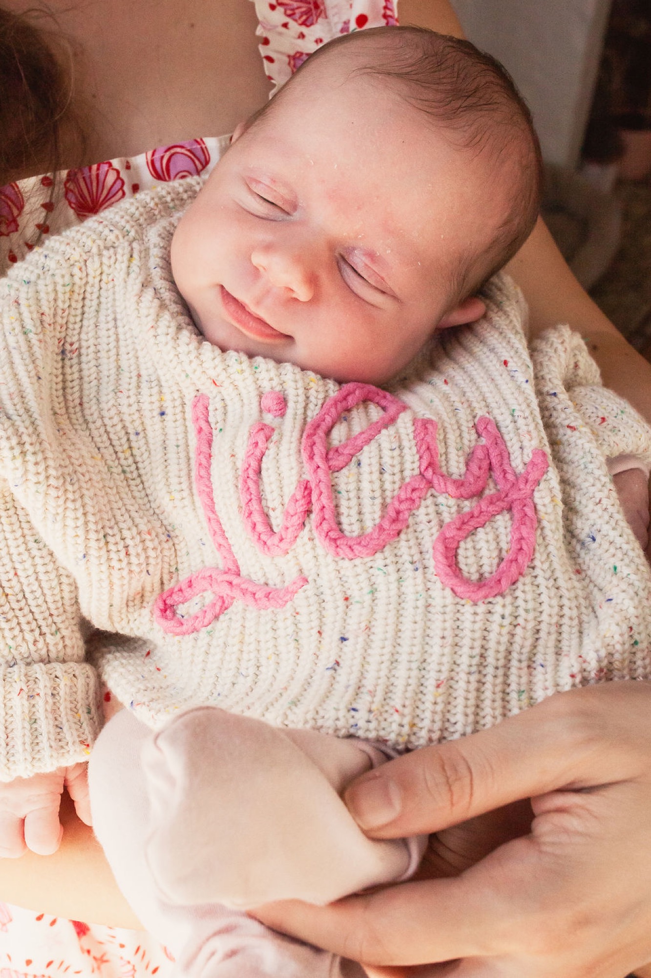 Newborn in cream knit sweater with pink lettering smiles peacefully while cradled in parent's hands during indoor session