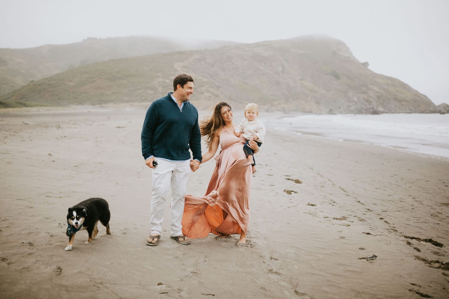 Pregnant mom holding toddler walks a foggy beach with dad and their dog, Marin County lifestyle photography