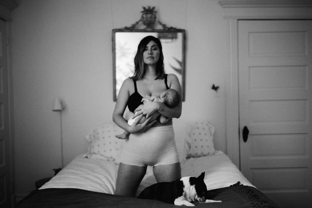 Black and white portrait of mom holding newborn on bed with Boston terrier nearby